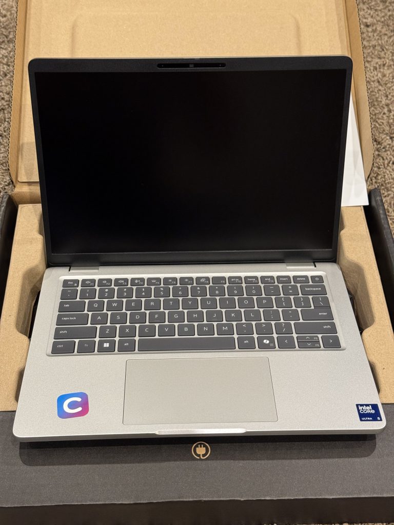 The silver Dell Pro 13 Plus laptop sitting in its open box, showing the keyboard, trackpad, and Intel Core Ultra sticker on the palm rest.